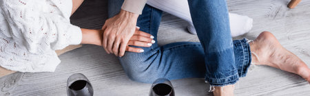 Cropped view of couple holding hands near glasses of wine on floor, banner