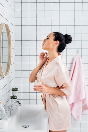 Young woman in silk bathrobe applying cosmetic serum near mirror in bathroomの写真素材
