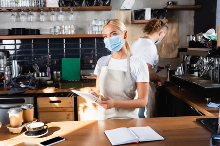 blonde waiter in medical mask holding digital tablet near notebook, coffee to go, and barista working on backgroundの写真素材