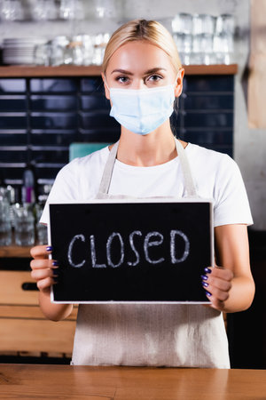 blonde barista in medical mask holding board with closed lettering in cafe