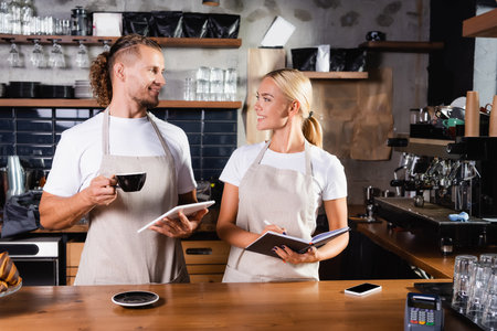 smiling barista with coffee cup and digital tablet near waiter with notebook, and payment terminal on bar counterの写真素材