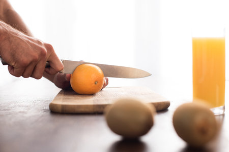 Cropped view of man cutting orange near kiwi and glass of orange juice on blurred foregroundの写真素材