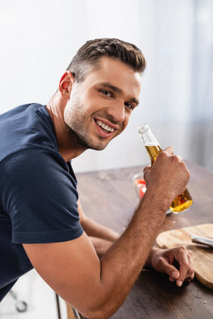 Smiling man holding bottle of beer near cutting board on blurred background in kitchenの写真素材