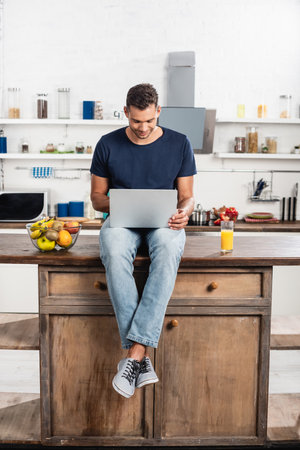 Young man using laptop near orange juice and fruits on kitchen tableの写真素材