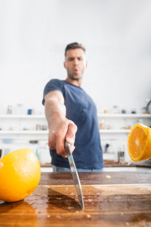 Close up view of halves of orange on wet cutting board and knife in hand of young man on blurred backgroundの写真素材