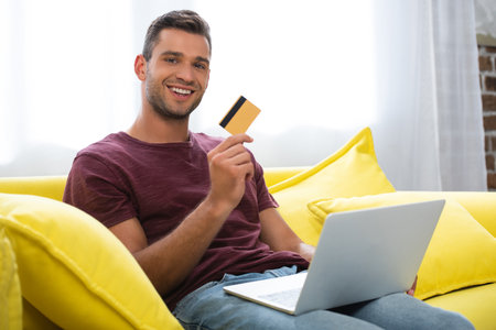 Cheerful man holding credit card and laptop on blurred foreground at homeの写真素材