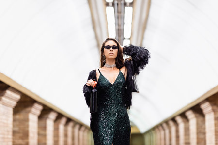 trendy woman in black lurex dress and sunglasses posing at underground station with bottle of wine