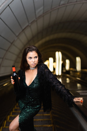 elegant young woman in black lurex dress holding wine bottle while looking away on escalator