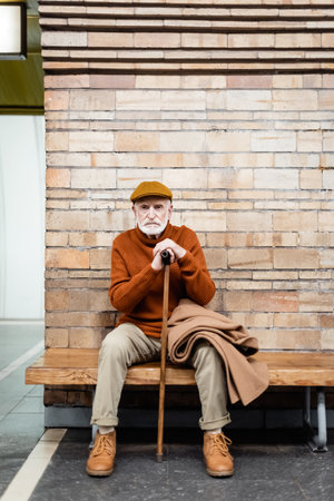 elderly man in autumn outfit sitting with walking stick on subway platform benchの写真素材
