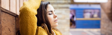 young woman in autumn clothes standing with closed eyes and hand on head near wall at metro station, bannerの写真素材