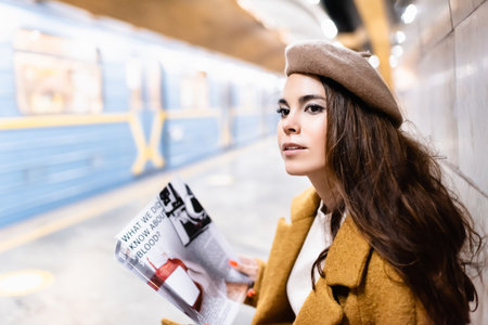 young woman in beret holding magazine while sitting on metro platform with blurred trainの写真素材