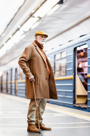 elderly man in cap and coat standing on metro station platform near train on blurred backgroundの写真素材