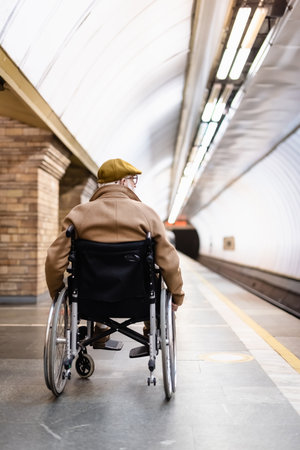 back view of senior man in wheelchair, wearing coat and cap, on platform of metro stationの写真素材
