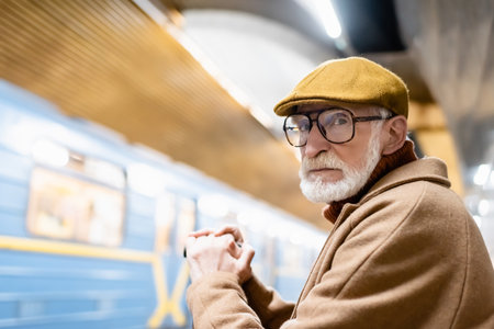 senior man in autumn clothes and eyeglasses looking at camera on metro platform with blurred trainの写真素材