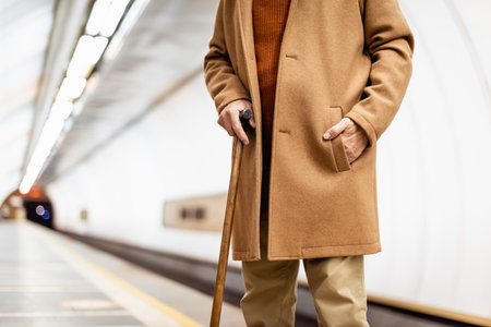 cropped view of aged man with walking stick, wearing autumn coat, standing on metro platformの写真素材