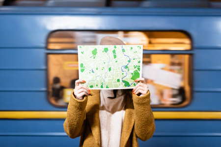 woman in autumn coat obscuring face with city map with blurred metro train on backgroundの写真素材