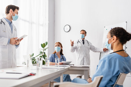 Doctor in medical mask pointing with finger near multiethnic colleagues with papers and digital tablet in clinicの写真素材