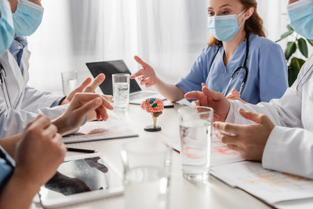 Doctors and nurses in medical masks talking while sitting at workplace with devices, pictures and glasses of water on blurred foregroundの写真素材
