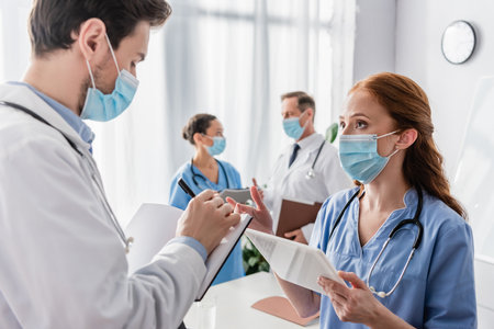 Redhead nurse talking to doctor writing in notebook with blurred multicultural colleagues on backgroundの写真素材