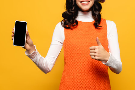 Cropped view of brunette woman with thumb up showing smartphone with blank screen isolated on yellow