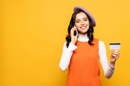 Smiling brunette woman in beret talking on smartphone and looking at camera while showing credit card isolated on yellowの写真素材