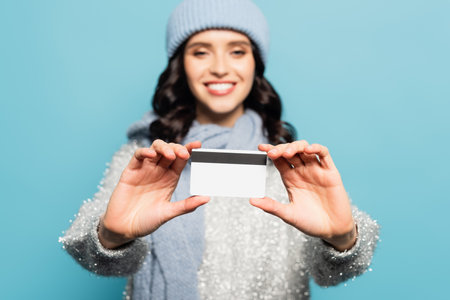 Cheerful brunette woman in winter outfit showing credit card on blurred background isolated on blueの写真素材