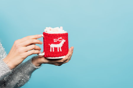 Cropped view of woman holding cup with marshmallows and knitted holder isolated on blueの写真素材
