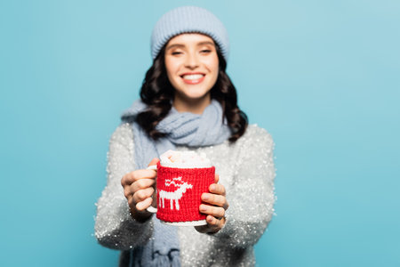 Cup with marshmallows and knitted holder in hands of blurred woman on background isolated on blue