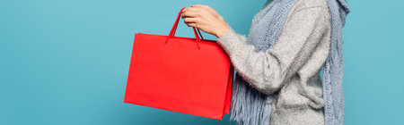 Cropped view of woman wearing scarf and sweater, holding red shopping bags isolated on blue, bannerの写真素材