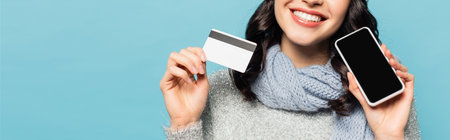 Cropped view of woman wearing scarf holding credit card and smartphone with blank screen isolated on blue, bannerの写真素材