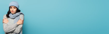 Displeased young adult woman in scarf and hat looking at camera while hugging herself isolated on blue, bannerの写真素材