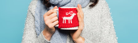 Cropped view of woman in scarf and pullover holding cup with knitted holder isolated on blue, bannerの写真素材