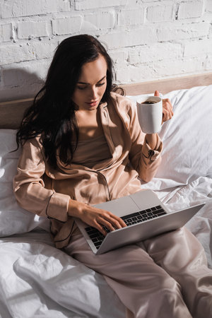 young brunette woman sitting in bed with mug and laptop at morningの写真素材