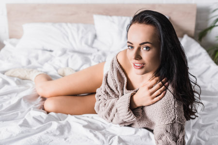 young brunette woman in sweater and socks lying in bed at morningの写真素材