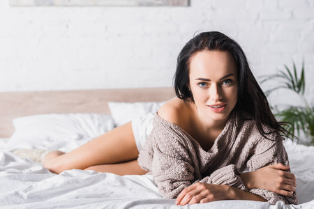 young brunette woman in sweater and socks lying in bed at morningの写真素材