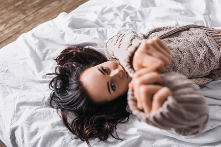 top view of young brunette woman in sweater and blurred outstretched hands lying in bed at morningの写真素材