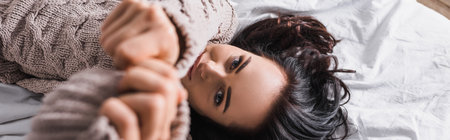 top view of young brunette woman in sweater and blurred outstretched hands lying in bed at morning, bannerの写真素材