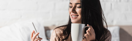 happy young brunette woman sitting in bed with mug and smartphone at morning, bannerの写真素材