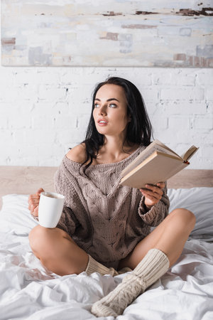 dreamy young brunette woman in sweater sitting in bed with mug of hot cocoa and book at morningの写真素材