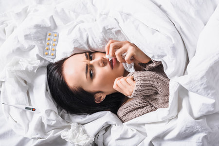 top view of sick young brunette woman with tissue, pills and thermometer in bedの写真素材