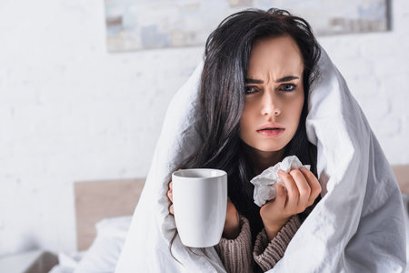 sick young brunette woman with tissue and hot drink in bedの写真素材