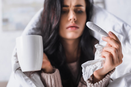sick young brunette woman with tissue, thermometer and hot drink in bed