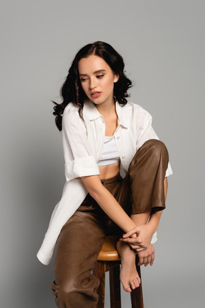 Curly brunette woman in casual clothes looking away while posing on stool on grayの写真素材