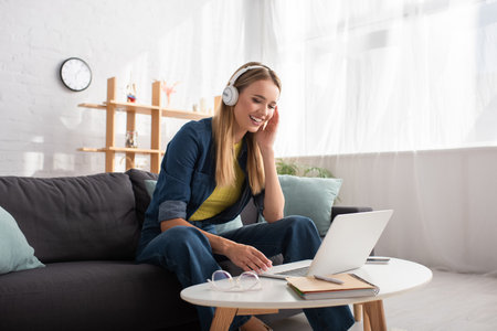 Cheerful young blonde woman in headphones looking at laptop while sitting on couch at homeの写真素材
