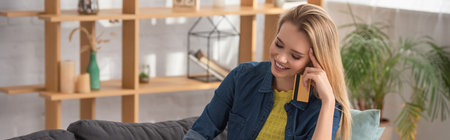 Cheerful blonde woman with credit card looking down at home on blurred background, bannerの写真素材
