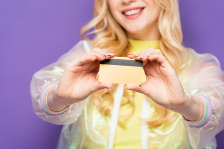 cropped view of happy blonde young woman in colorful outfit showing credit card on purple background
