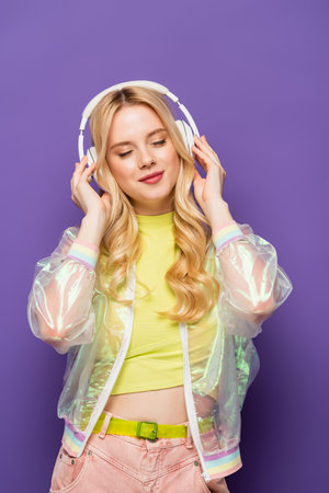 blonde young woman in colorful outfit and headphones with closed eyes on purple backgroundの写真素材