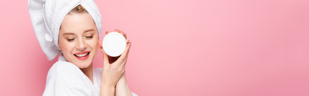 happy young woman in bathrobe with closed eyes, towel on head holding cosmetic cream isolated on pink, bannerの写真素材