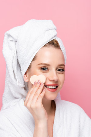 happy young woman in bathrobe with towel on head and facial heart shaped sponge isolated on pinkの写真素材