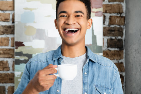 amazed african american man laughing while holding cup of coffeeの写真素材